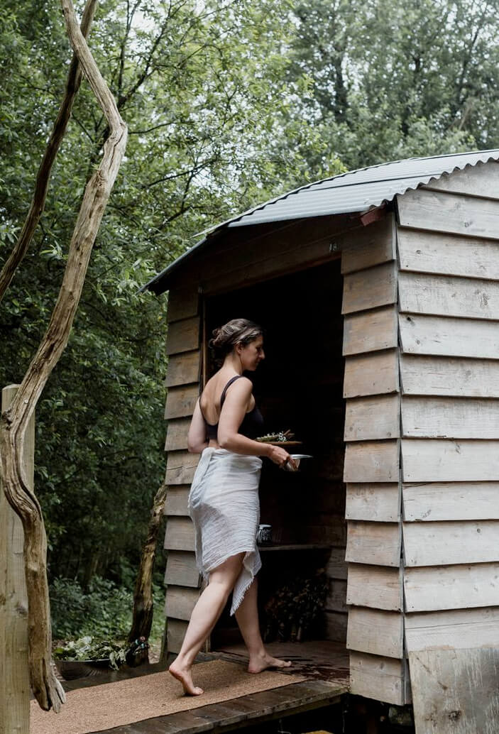 a woman dressed in a bathing suit and towel around her hip walking into a shed looking sauna situated in the woods