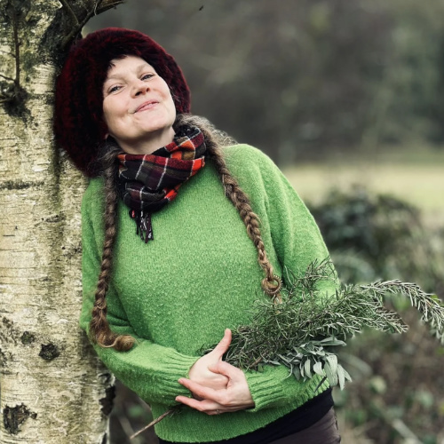 A smiling woman wearing a green whooly jumper is leaning against a tree holding a few tree branches